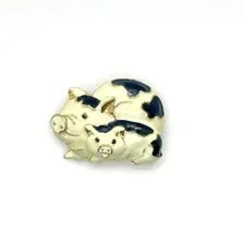 A whimsical vintage gold-tone brooch featuring a stacked pair of cream enamel pigs decorated with smooth black cow-print spots. The playful design highlights polished gold-plated snouts, ears, and hooves, cleanly photographed against a neutral light gray background.