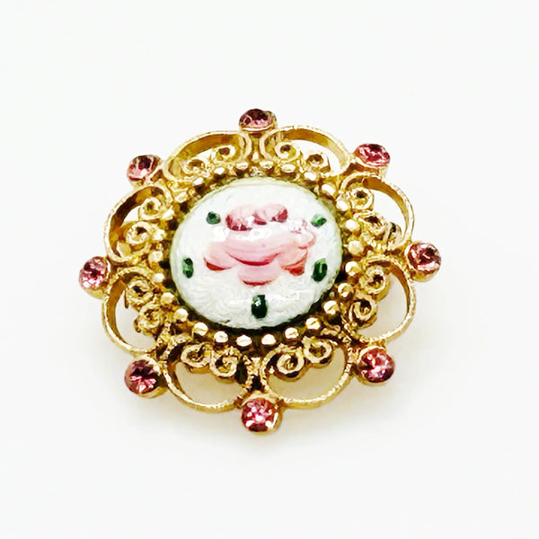 Take a look at this vintage costume jewelry brooch featuring a hand-painted pink rose on a white enamel or ceramic base, set within an ornate gold-tone filigree frame accented with pink rhinestones. The center showcases a pink rose, with green dots accenting the perimeter. The setting and metalwork around the outside are particularly lovely. It measures 1" across.