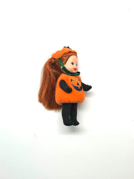 Vintage Mattel Kelly Club 2000 Halloween Party Pumpkin Jenny doll. Small doll with long brown hair wearing an orange plush pumpkin costume with a printed jack-o’-lantern face, black long sleeves, black pants, and a matching orange pumpkin cap with a green strap on a white background