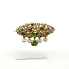 Product photo of a vintage antique-gold filigree pin. The central oval frame holds bright green and red glass gems. Six small dangling charms hang from the bottom, including five creamy white pearls and one round green rhinestone.