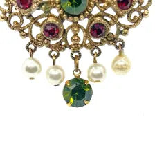 Product photo of a vintage antique-gold filigree pin. The central oval frame holds bright green and red glass gems. Six small dangling charms hang from the bottom, including five creamy white pearls and one round green rhinestone.