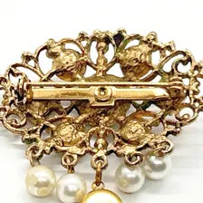 Product photo of a vintage antique-gold filigree pin. The central oval frame holds bright green and red glass gems. Six small dangling charms hang from the bottom, including five creamy white pearls and one round green rhinestone.