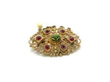 Product photo of a vintage antique-gold filigree pin. The central oval frame holds bright green and red glass gems. Six small dangling charms hang from the bottom, including five creamy white pearls and one round green rhinestone.