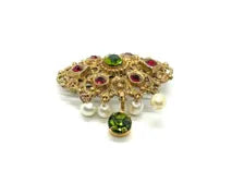 Product photo of a vintage antique-gold filigree pin. The central oval frame holds bright green and red glass gems. Six small dangling charms hang from the bottom, including five creamy white pearls and one round green rhinestone.