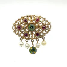 Product photo of a vintage antique-gold filigree pin. The central oval frame holds bright green and red glass gems. Six small dangling charms hang from the bottom, including five creamy white pearls and one round green rhinestone.