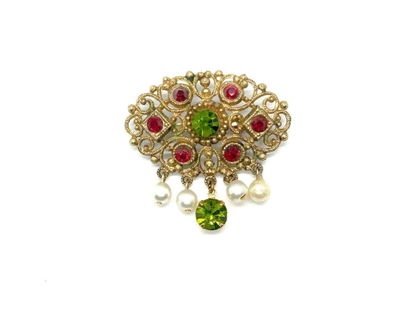 Product photo of a vintage antique-gold filigree pin. The central oval frame holds bright green and red glass gems. Six small dangling charms hang from the bottom, including five creamy white pearls and one round green rhinestone.