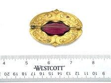 Antique Art Nouveau sash pin brooch crafted from embossed gold-tone brass with swirling whiplash foliage curves, featuring a large prong-set oval faceted purple amethyst glass gemstone center.