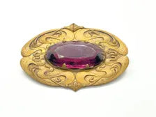 Antique Art Nouveau sash pin brooch crafted from embossed gold-tone brass with swirling whiplash foliage curves, featuring a large prong-set oval faceted purple amethyst glass gemstone center.