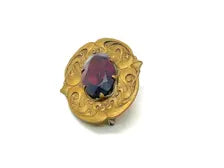Antique Art Nouveau sash pin brooch crafted from embossed gold-tone brass with swirling whiplash foliage curves, featuring a large prong-set oval faceted purple amethyst glass gemstone center.