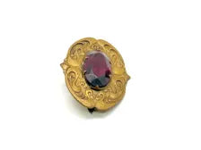Antique Art Nouveau sash pin brooch crafted from embossed gold-tone brass with swirling whiplash foliage curves, featuring a large prong-set oval faceted purple amethyst glass gemstone center.