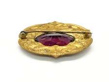 An ornate, gold-toned antique brooch viewed from its reverse side, featuring a large, oval-faceted purple gemstone or glass center housed within an embossed, decorative metallic frame. A long, thin horizontal safety pin with a classic C-clasp fastener stretches across the upper back of the piece.