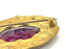 An extreme close-up, side-angle view of an antique Art Nouveau gold-tone sash brooch pin resting on a plain white surface. The brooch features an ornate, embossed metallic frame with swirling, organic patterns. Set deeply inside the frame is a large, faceted purple rhinestone or glass gem. A long, thin metal pin stem stretches horizontally across the upper portion of the frame, secured into a simple "C" style catch on the right side.