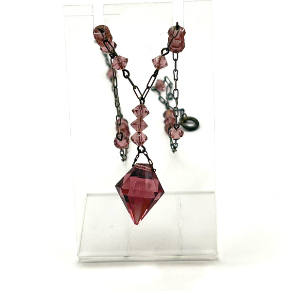 A vintage necklace featuring clusters of deep purple bicone crystal beads on a dark-toned paperclip chain, culminating in a large, heavily faceted diamond-shaped purple crystal drop pendant, displayed on a white jewelry bust