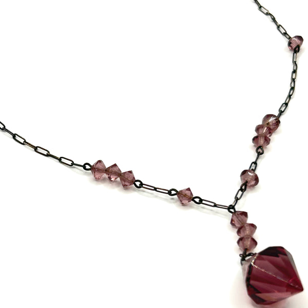 A vintage necklace featuring clusters of deep purple bicone crystal beads on a dark-toned paperclip chain, culminating in a large, heavily faceted diamond-shaped purple crystal drop pendant
