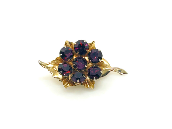 A vintage-style gold flower brooch featuring five faceted dark purple gemstones, set against a plain white background.