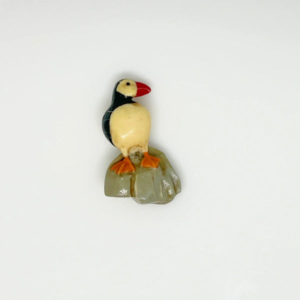 This is a brooch by artist Ralph Massey, who is acclaimed for many types of artwork- acrylic, performance, resin and more. This brooch is resin, hand made and hand painted in white, black, red-orange and gray. It depicts a penguin standing on rock, and measures about 1.75” inches tall.