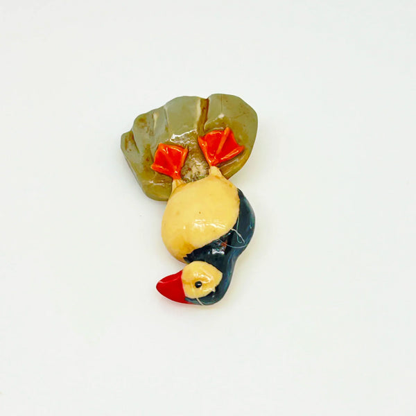 This is a brooch by artist Ralph Massey, who is acclaimed for many types of artwork- acrylic, performance, resin and more. This brooch is resin, hand made and hand painted in white, black, red-orange and gray. It depicts a penguin standing on rock, and measures about 1.75” inches tall.