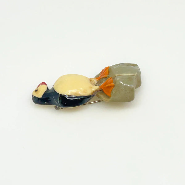This is a brooch by artist Ralph Massey, who is acclaimed for many types of artwork- acrylic, performance, resin and more. This brooch is resin, hand made and hand painted in white, black, red-orange and gray. It depicts a penguin standing on rock, and measures about 1.75” inches tall.