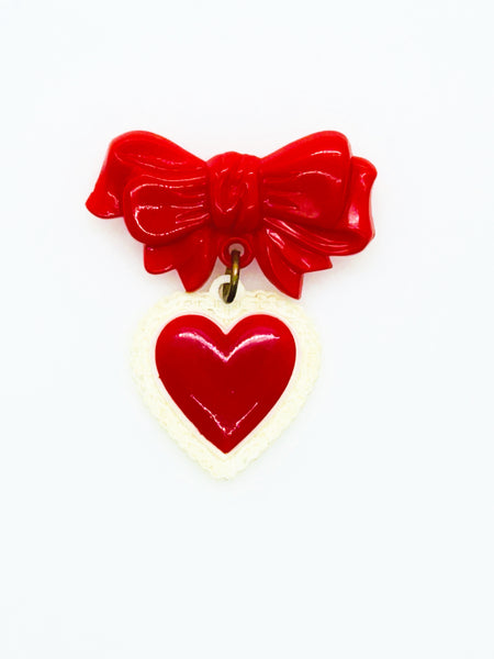 This lovely and nostalgic piece is a vintage early plastic sweetheart bow with dangling filigree lace heart brooch pin, often described as a "Heart Within a Heart" design. It is a recognized "book piece" in vintage jewelry collecting and was commonly made from materials like celluloid plastic .