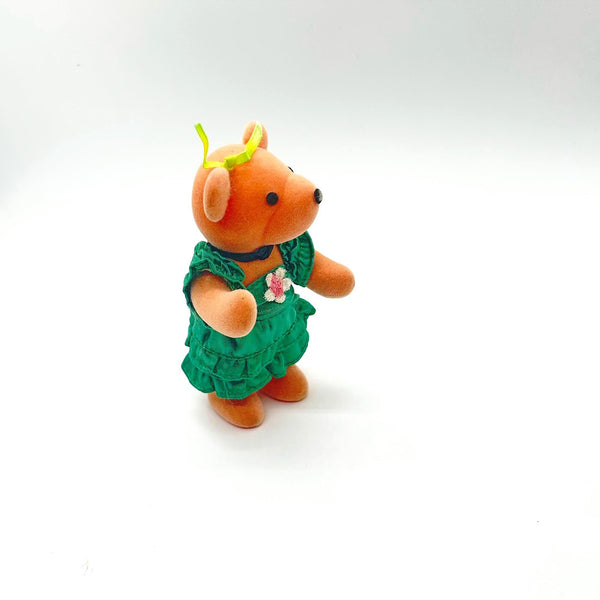 A small plush teddy bear ornament wearing a green floral dress and a yellow bow, standing against a plain white background.