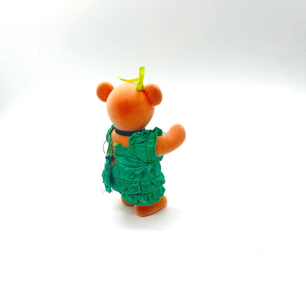 A small orange plush bear figurine shown from the back, wearing a ruffled green dress and a small yellow bow on its head, photographed against a plain white background.