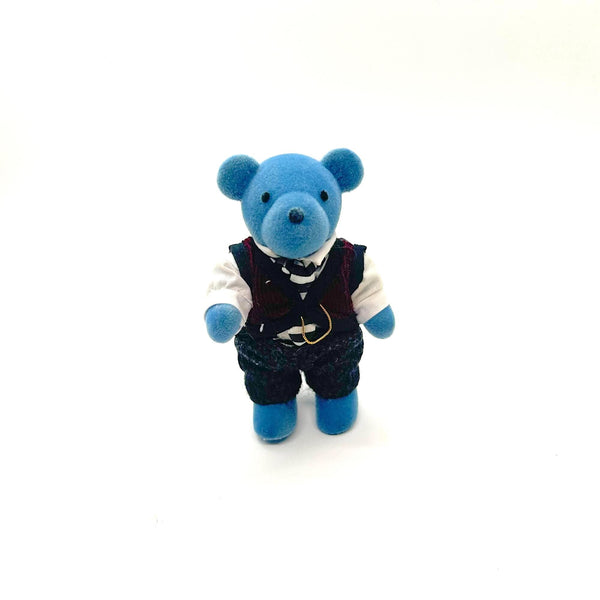 A small blue teddy bear figurine wearing a white shirt, dark vest, striped tie, and dark trousers, standing against a plain white background.