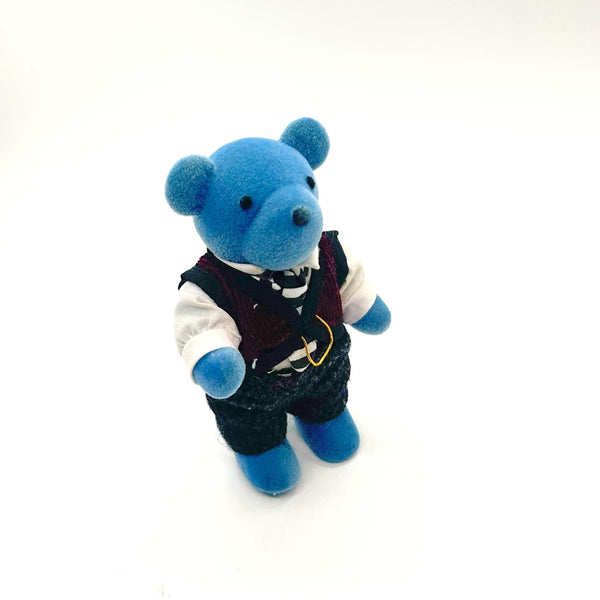 A small blue teddy bear figurine wearing a white shirt, dark vest, striped tie, and dark trousers, standing against a plain white background.