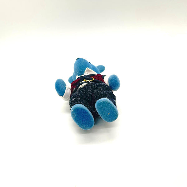 A small blue plush toy figure lying on its back, wearing a dark textured outfit with a red collar detail, photographed on a plain white background.