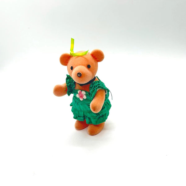 A small plush teddy bear ornament wearing a green floral dress and a yellow bow, standing against a plain white background.