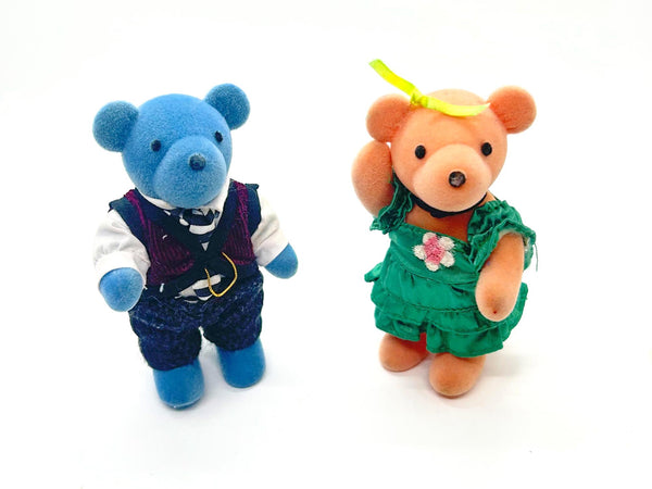 Two small plush teddy bears standing side by side on a white background. The left bear is blue and wears a white shirt, dark vest, and dark pants.