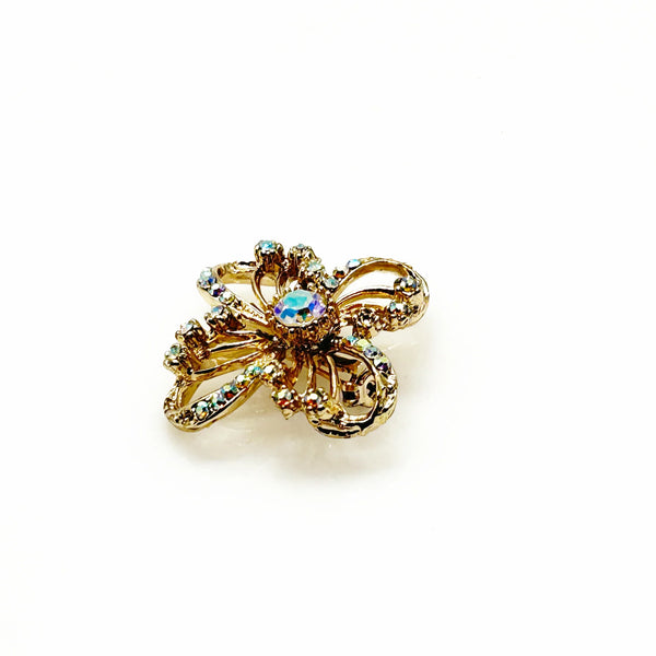 This is a three dimensional vintage ribbon brooch featuring clear rhinestones and a central aurora borealis (AB) gemstone, designed in an abstract floral or bow shape. 