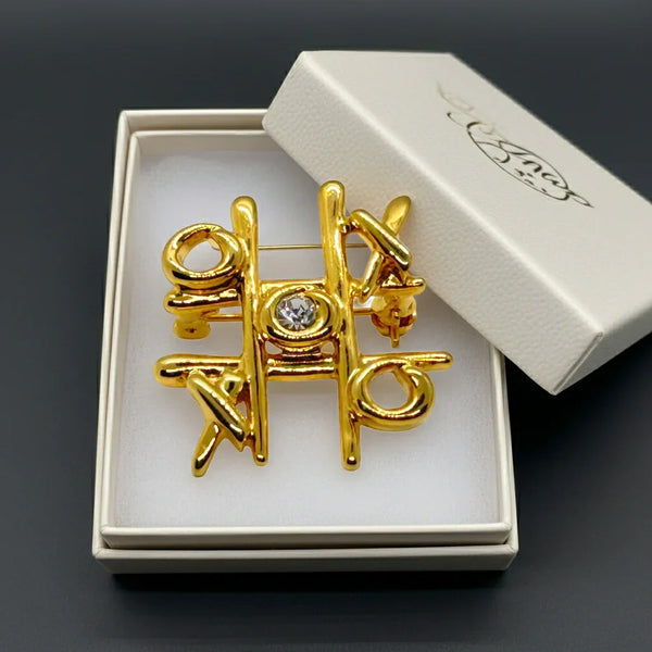 This charming Anne Klein brooch captures the playful spirit of vintage costume jewelry with its clever tic-tac-toe design. Crafted in warm gold tone, the piece features an open-style board complete with X's and O's, crowned by a sparkling clear rhinestone at its center