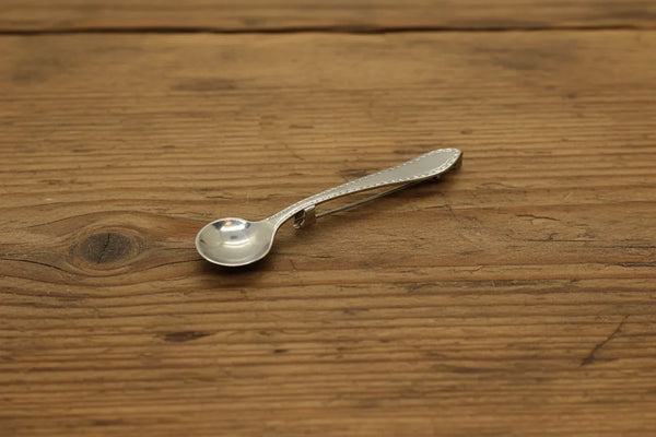 A vintage silver-toned metal pin with a spoon-shaped head and a long, narrow pin stem, photographed on a wood background