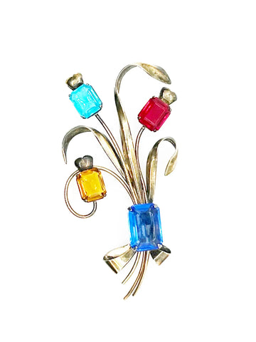 Experience the charm and beauty of this stunning vintage multicolored brooch. It features a vibrant and stylized flower with emerald cut stones in citrine, aqua, red, and blue. Near the base is larger, deep blue stone
