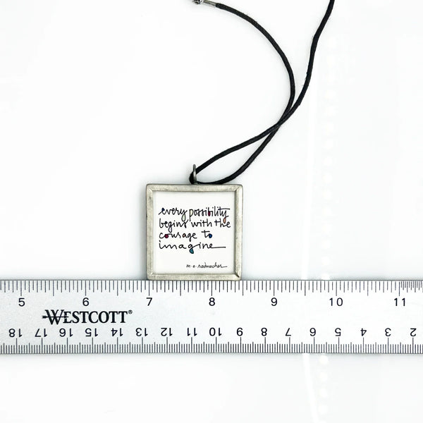 This is a modern inspiration in the form of a square pendant centering around the quote "Every possibility begins with the courage to imagine" by Mary Anne Radmacher. The quote is written in black ink, on a white background, and framed in silver metal. The reverse features a blue night sky background with a quarter moon and star. 