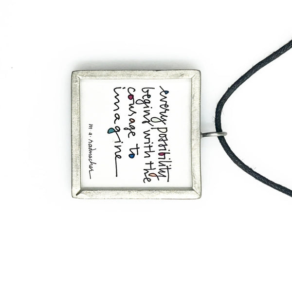 This is a modern inspiration in the form of a square pendant centering around the quote "Every possibility begins with the courage to imagine" by Mary Anne Radmacher. The quote is written in black ink, on a white background, and framed in silver metal. The reverse features a blue night sky background with a quarter moon and star. 