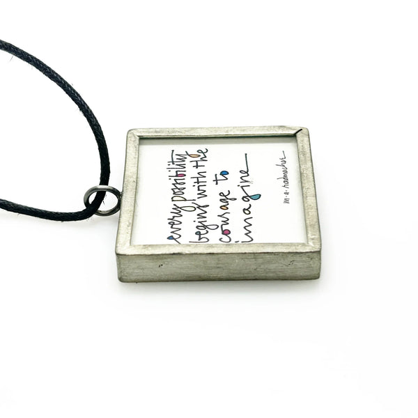This is a modern inspiration in the form of a square pendant centering around the quote "Every possibility begins with the courage to imagine" by Mary Anne Radmacher. The quote is written in black ink, on a white background, and framed in silver metal. The reverse features a blue night sky background with a quarter moon and star. 