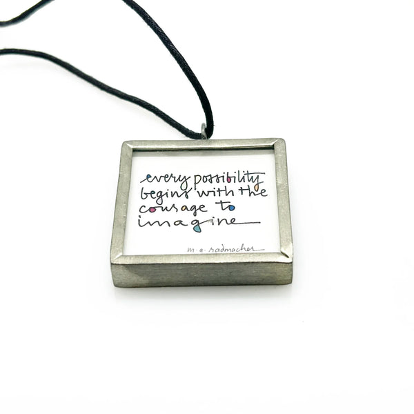 This is a modern inspiration in the form of a square pendant centering around the quote "Every possibility begins with the courage to imagine" by Mary Anne Radmacher. The quote is written in black ink, on a white background, and framed in silver metal. The reverse features a blue night sky background with a quarter moon and star. 