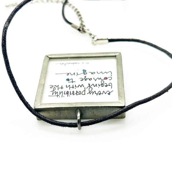 This is a modern inspiration in the form of a square pendant centering around the quote "Every possibility begins with the courage to imagine" by Mary Anne Radmacher. The quote is written in black ink, on a white background, and framed in silver metal. The reverse features a blue night sky background with a quarter moon and star. 