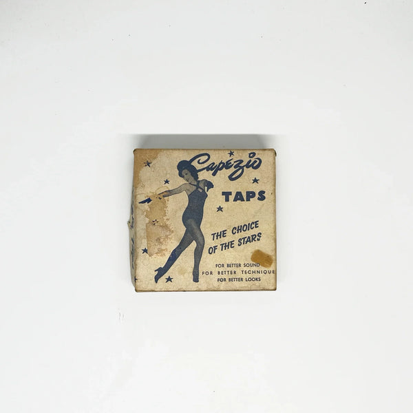 The square box measures 3.25: wide by 3.25" tall. The front shows a photo of a spirited female tap dancer. Inside the box, there are two taps, each engraved Capezio, and a small packet containing the nails or grips. They were used on Capezio character shoes and K360 oxfords.