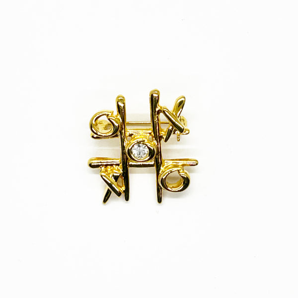 This charming Anne Klein brooch captures the playful spirit of vintage costume jewelry with its clever tic-tac-toe design. Crafted in warm gold tone, the piece features an open-style board complete with X's and O's, crowned by a sparkling clear rhinestone at its center