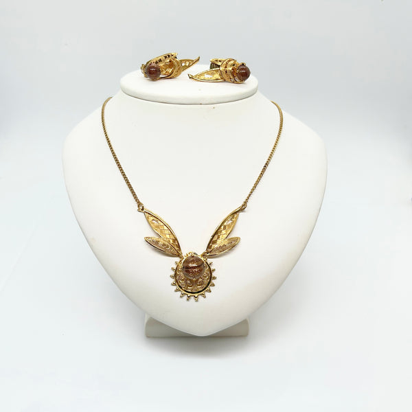 A vintage German jewelry parure set featuring a gold-tone filigree necklace and matching clip-on earrings displayed on a white neck bust. The pendant and earrings showcase a winged leaf motif set with amber-colored goldstone art glass cabochons.