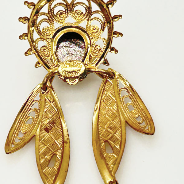 A close-up of the back ornate gold-toned jewelry piece, likely a brooch or pendant, featuring a circular filigree top, a central oval inset with a textured silver-toned center, and two dangling leaf-shaped filigree pendants below, shown against a plain light background.