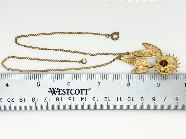 A gold-toned chain necklace with a pendant featuring leaf-like elements and a sunburst design, shown above a white ruler for scale. The ruler has the brand name WESTCOTT printed on it.