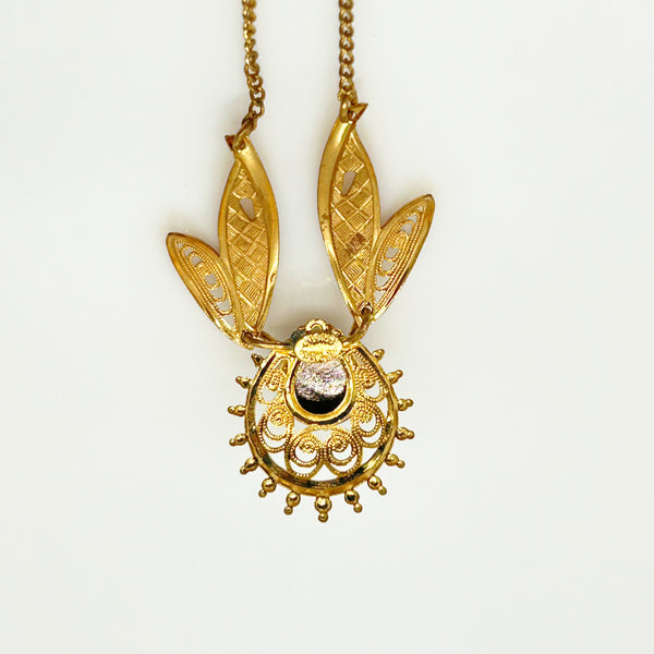 A gold pendant necklace featuring a stylized sun or beetle-like design with two leaf-shaped wings, intricate filigree detailing, and a central dark oval stone, shown against a plain white background.