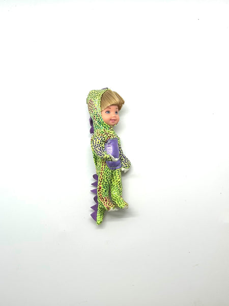A vintage 2003 Mattel Kelly Club 4-inch Tommy doll dressed in a green dragon Halloween costume with a purple chest patch and purple spiked tail. The mini pocket doll has blonde hair and blue eyes, photographed standing against a solid white background