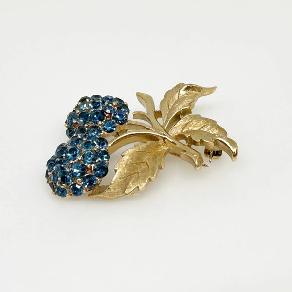 This vintage Crown Trifari brooch features a design of two berries suspended from intricate leaves and stems. The berries boast a captivating medium blue hue, beautifully set in a crafted gold-tone setting that gives the appearance of an invisible or pave setting