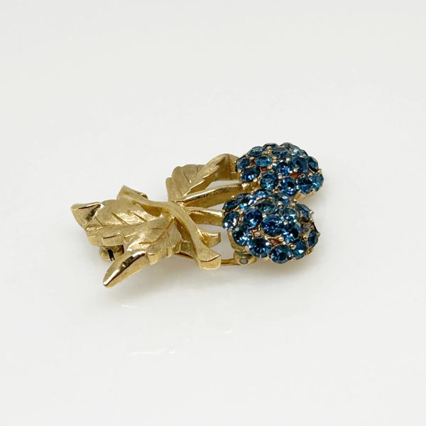 This vintage Crown Trifari brooch features a design of two berries suspended from intricate leaves and stems. The berries boast a captivating medium blue hue, beautifully set in a crafted gold-tone setting that gives the appearance of an invisible or pave setting