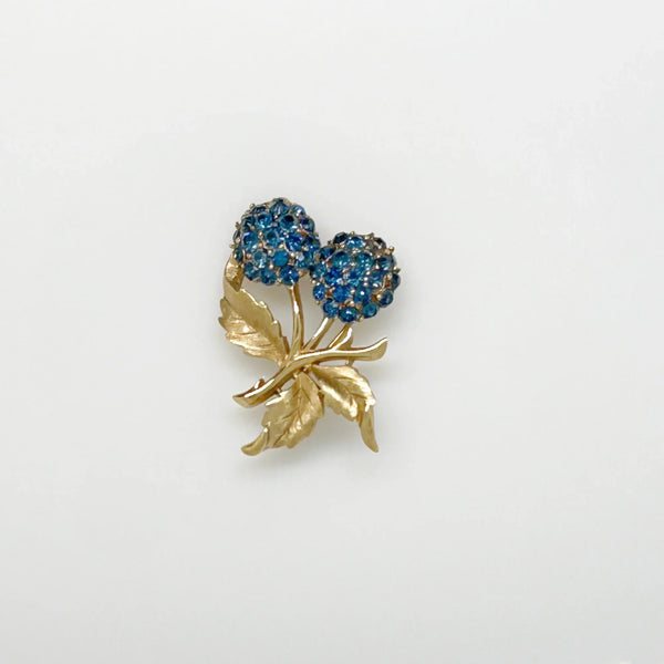 This vintage Crown Trifari brooch features a design of two berries suspended from intricate leaves and stems. The berries boast a captivating medium blue hue, beautifully set in a crafted gold-tone setting that gives the appearance of an invisible or pave setting