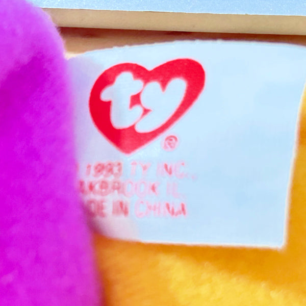Close-up of a Ty brand fabric tag sewn onto a plush toy, showing the red heart logo and 'ty' text on a white label, with purple and yellow plush material visible around it.